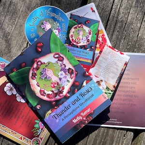 May include: A collection of printed materials featuring floral and pomegranate designs. The items include a round sticker, cards with text, and a booklet titled "Thunder and Roses: A Persephone Devotional" by Molly Remer.