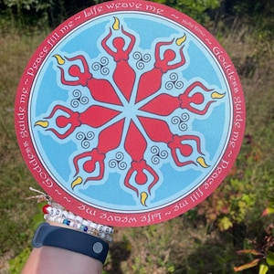 May include: A round, colorful art piece featuring a central design of red figures with raised arms, radiating from a center. The figures are set against a light blue background. The outer ring has text that reads "Goddess guide me ~ Peace fill me ~ Life weave me".