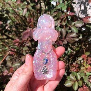 May include: A pink resin figurine of a woman with iridescent glitter embedded in the resin. The figurine has a silver chain necklace with a blue and white bead and a silver flower charm.