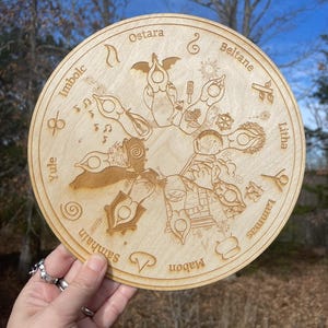 May include: A round wooden decorative piece with engraved symbols and text. The text includes the words "Imbolc", "Ostara", "Beltane", "Litha", "Lammas", "Mabon", "Samhain", and "Yule". The piece is held in a hand.