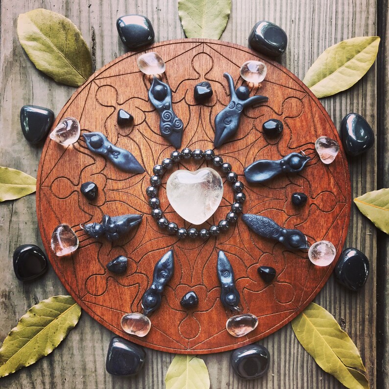 May include: A wooden circle with a carved design, surrounded by black stones and crystals. In the centre is a clear quartz heart, surrounded by a beaded necklace. There are also small black figurines of women around the circle.
