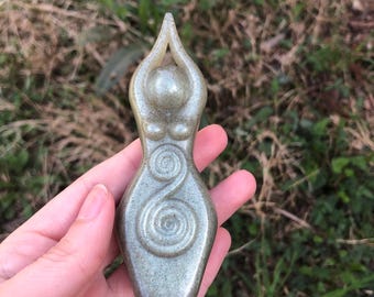 May include: A light green stone figurine of a female form with arms raised above the head. The figure has a rounded head, breasts, and a spiral design on the torso. The statue is held in a hand against a background of green grass.
