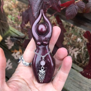 May include: A dark purple wooden figurine of a woman with her arms raised above her head. The figurine has a silver chain necklace with a pink bead and a silver owl charm.