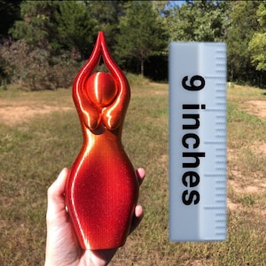 May include: A red and orange figurine of a female form with arms raised. The sculpture is approximately 9 inches tall. The background is a grassy field with trees.