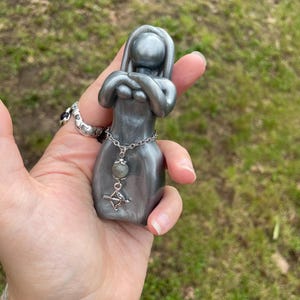 May include: A gray figurine of a woman with arms crossed, adorned with a silver chain and a small charm. The figurine is held in a hand against a blurred green background. The charm features a bow and arrow.