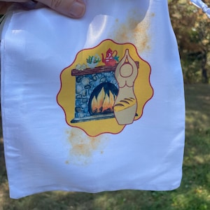 Puede incluir: Una bolsa blanca con cordón que presenta un gráfico central de una chimenea de piedra con fuego, una tetera roja y una figura estilizada sosteniendo un pan, sobre un fondo amarillo festoneado con manchas naranjas en la tela.