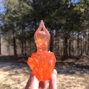 May include: A translucent orange resin figurine of a female form with arms raised above the head. The figure is atop a cluster of orange crystal-like formations. The background is a blurred view of trees and sunlight.