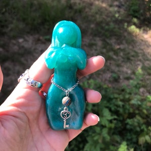 Puede incluir: Una figurita de resina de color verde azulado de una mujer con un collar de plata con un dije de llave. La figurita tiene un acabado brillante y está sosteniendo sus manos juntas frente a su pecho.