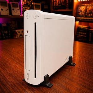 Könnte beinhalten: Eine weiße Wii U Spielkonsole, die vertikal auf zwei schwarzen Kunststoffständern steht. Die Vorderseite verfügt über einen Power-Knopf, eine Auswurftaste, einen Disc-Schlitz, eine Sync-Taste und das Wii U Logo in schwarzer Schrift.