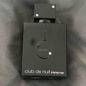 Könnte beinhalten: Eine schwarze, rechteckige Parfümflasche mit quadratischem Verschluss. Auf der Flasche steht in Weiß der Text "club de nuit intense man". Eine kleine Kette ist an einem runden Anhänger mit dem Markennamen "ARMAF" befestigt. Der Verschluss und der Anhänger haben dekorative Strasssteine.