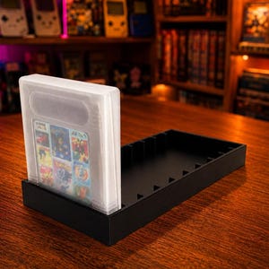 Könnte beinhalten: Ein schwarzes Kunststoff-Aufbewahrungsfach für Handheld-Videospielkassetten mit mehreren vertikalen Schlitzen zur Organisation. Eine durchsichtige Schutzhülle, die eine Spielkassette mit buntem Etikett enthält, ist in den ersten Schlitz eingesetzt.