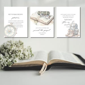 Könnte beinhalten: Drei Aquarelldrucke mit religiösen Themen. Einer zeigt einen Kompass, ein anderer ein offenes Buch mit Brille und der dritte einen Rucksack und Stiefel. Die Drucke haben Texte aus der Bibel. Ein offenes Buch mit einem Band ist im Vordergrund.
