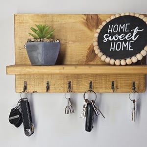 Può includere: Porta chiavi in legno con uno scaffale e sei ganci. Il porta chiavi ha un cartello che dice "Home Sweet Home" e una piccola pianta grassa in vaso.