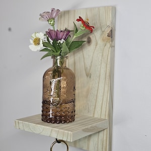 Puede incluir: Una estantería de pared de madera con un pequeño estante y un gancho. Un jarrón de vidrio marrón con un patrón floral contiene un ramo de flores rosas, blancas y rojas.