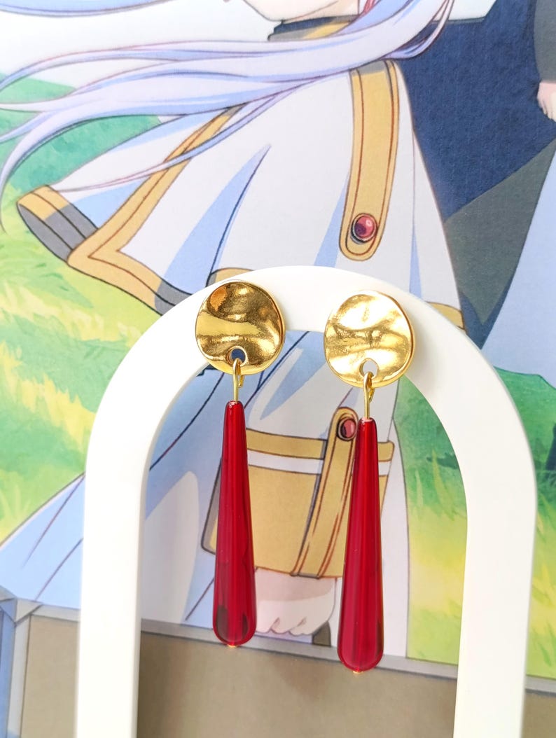 Puede incluir: Pendientes dorados con un dise&ntilde;o de disco martillado y largos colgantes rojos en forma de l&aacute;grima. Los pendientes se exhiben en un soporte blanco sobre un fondo de una ilustraci&oacute;n de estilo anime.