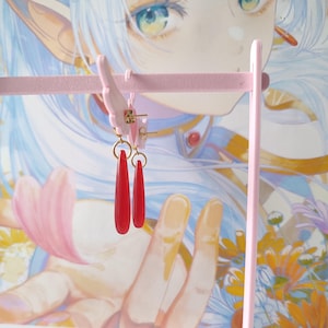 Puede incluir: Un par de pendientes rojos en forma de l&aacute;grima colgados de un soporte rosa para pendientes. Los pendientes tienen un aro dorado y un colgante largo y delgado de color rojo. El soporte para pendientes es rosa y tiene un dise&ntilde;o decorativo.