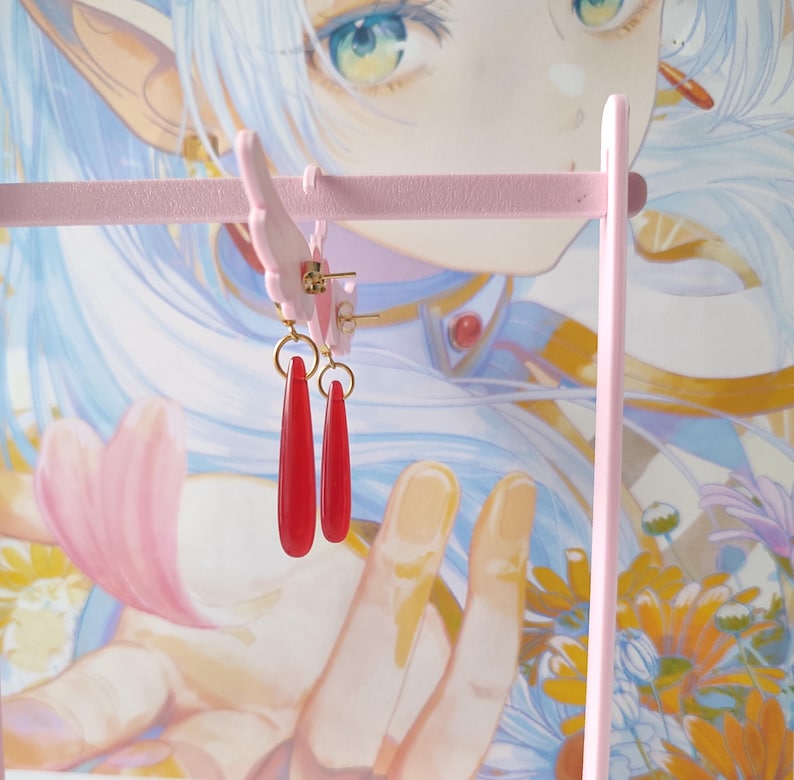 Puede incluir: Un par de pendientes rojos en forma de l&aacute;grima colgados de un soporte rosa para pendientes. Los pendientes tienen un aro dorado y un colgante largo y delgado de color rojo. El soporte para pendientes es rosa y tiene un dise&ntilde;o decorativo.
