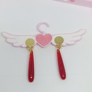 Puede incluir: Un par de pendientes rojos en forma de l&aacute;grima con detalles dorados, colgados de un expositor rosa en forma de ala. El expositor tiene un coraz&oacute;n rosa brillante y la palabra "MAGICAL" en letras rosas.