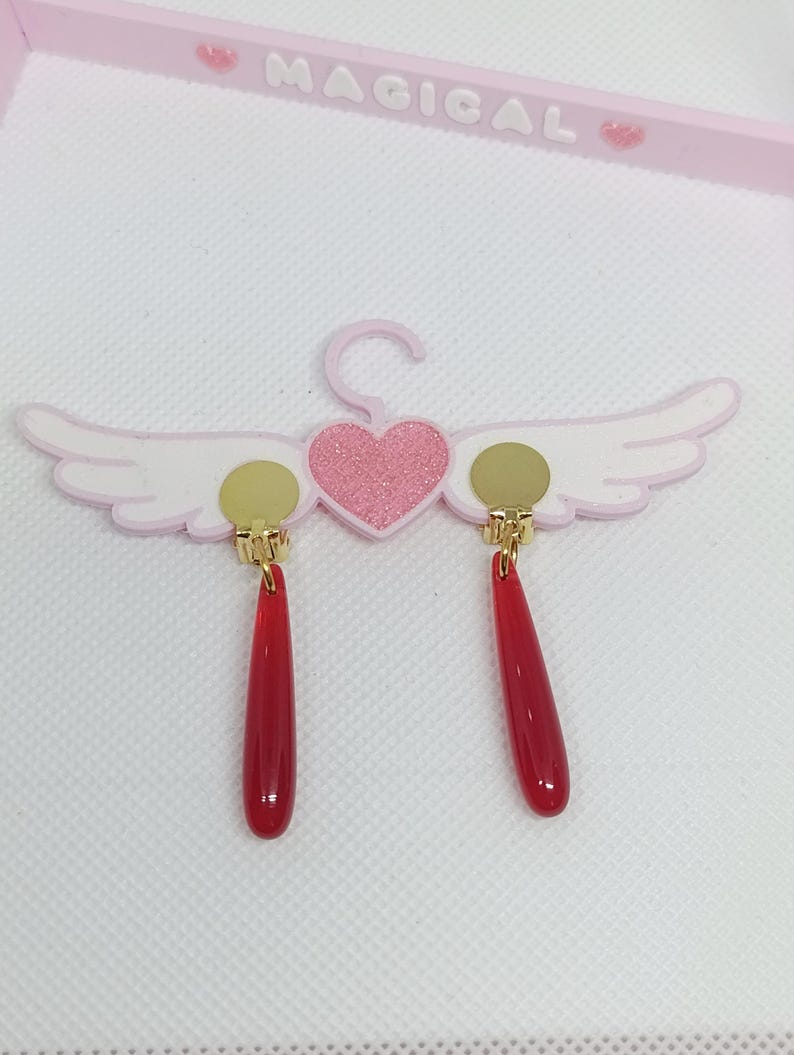 Puede incluir: Un par de pendientes rojos en forma de l&aacute;grima con detalles dorados, colgados de un expositor rosa en forma de ala. El expositor tiene un coraz&oacute;n rosa brillante y la palabra "MAGICAL" en letras rosas.