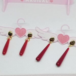 Puede incluir: Cuatro pares de pendientes rojos en forma de l&aacute;grima con tachuelas doradas. Los pendientes se exhiben en una percha decorativa rosa con detalles de coraz&oacute;n y alas. La palabra "MAGICAL" est&aacute; impresa en la parte superior de la percha.