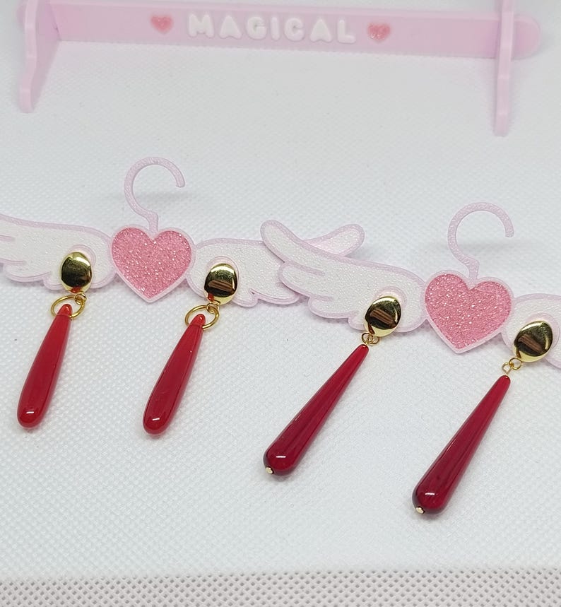 Puede incluir: Cuatro pares de pendientes rojos en forma de l&aacute;grima con tachuelas doradas. Los pendientes se exhiben en una percha decorativa rosa con detalles de coraz&oacute;n y alas. La palabra "MAGICAL" est&aacute; impresa en la parte superior de la percha.