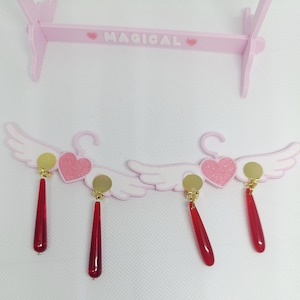 Puede incluir: Pendientes rosas y blancos con forma de alas de &aacute;ngel y colgantes rojos en forma de l&aacute;grima. Cada pendiente presenta un disco dorado y un coraz&oacute;n rosa con purpurina. Los pendientes se exhiben en un soporte de madera rosa con la palabra "MAGICAL".