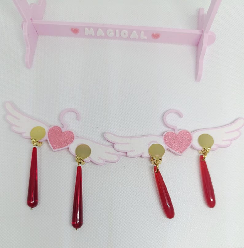 Puede incluir: Pendientes rosas y blancos con forma de alas de &aacute;ngel y colgantes rojos en forma de l&aacute;grima. Cada pendiente presenta un disco dorado y un coraz&oacute;n rosa con purpurina. Los pendientes se exhiben en un soporte de madera rosa con la palabra "MAGICAL".