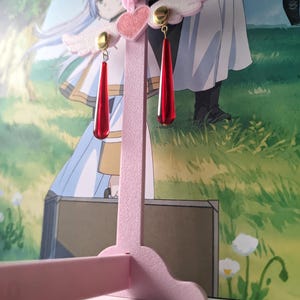 Puede incluir: Un expositor de pendientes rosa con dise&ntilde;o de coraz&oacute;n y alas, que sostiene dos pares de pendientes rojos en forma de l&aacute;grima con detalles dorados. Los pendientes cuelgan de postes dorados.