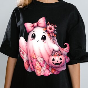Puede incluir: Camiseta negra con una ilustración de fantasma rosa. El fantasma tiene un lazo, flores y un cubo de calabaza. La parte inferior del fantasma tiene un diseño de patchwork. Un diseño caprichoso de Halloween.