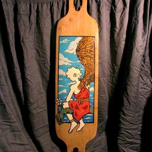 May include: A wooden longboard with a hand-painted design. The design features a cartoon character with a red dress and a yellow crown, sitting on a rock with a flower. The background is a blue sky with white clouds and a yellow field of wheat.