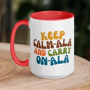 May include: White ceramic mug with a red rim. The mug has a colorful graphic that reads "Keep Calm-Ala and Carry On-Ala".