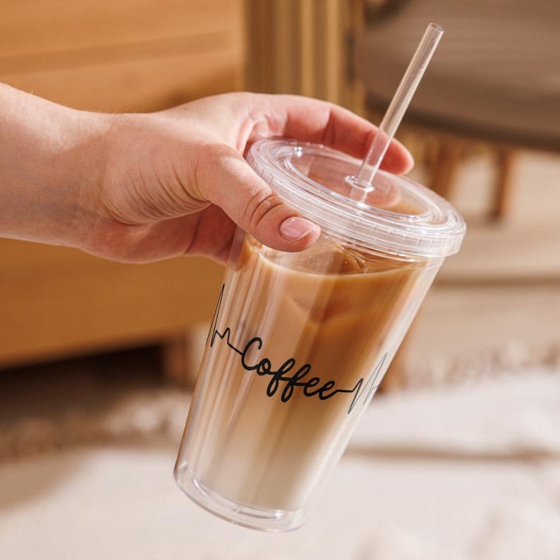 May include: A clear plastic tumbler with a black lid and a straw. The tumbler is filled with iced coffee and has the text "Coffee" written on it in a black cursive font.