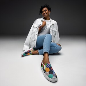May include: A person wearing a white denim jacket and blue jeans sits on a white surface. They are wearing a pair of colorful patchwork slip-on shoes.