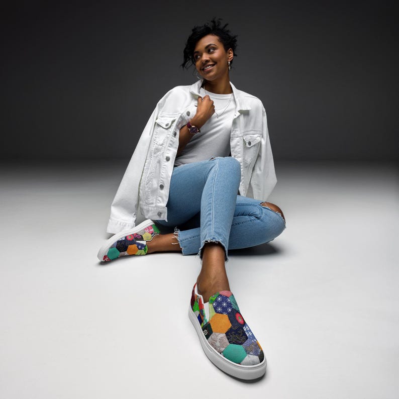 May include: A person wearing a white denim jacket and blue jeans sits on a white surface. They are wearing a pair of colorful patchwork slip-on shoes.