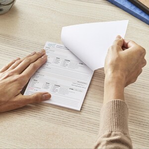 May include: A white notepad with printed text is being opened to reveal a blank page. The notepad has a light blue logo and the words "HYGIENE PATIENT HUDDLE PLANNER". The notepad is on a light wood surface.