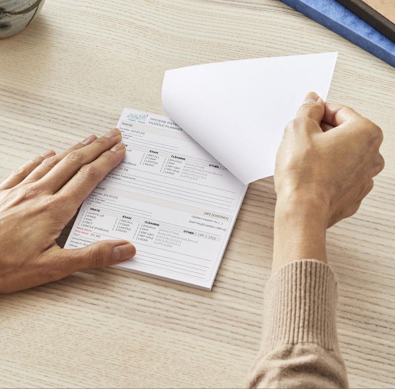 May include: A white notepad with printed text is being opened to reveal a blank page. The notepad has a light blue logo and the words "HYGIENE PATIENT HUDDLE PLANNER". The notepad is on a light wood surface.