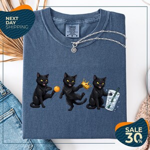a blue t-shirt with a graphic design of three black cats, each holding a different object: a ball, a crown, and a glass of water. The cats are depicted in a playful and whimsical manner, with one cat holding a crown and the other two holding a ball and a glass of water. The t-shirt is displayed on a white background, and there are some jeans and a beige sweater visible in the background.