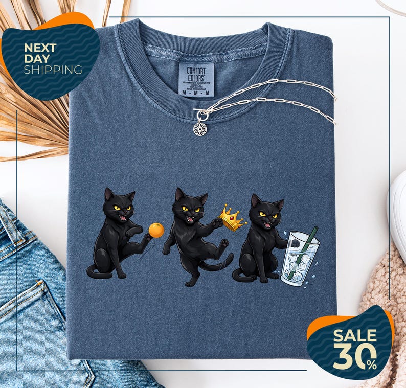 a blue t-shirt with a graphic design of three black cats, each holding a different object: a ball, a crown, and a glass of water. The cats are depicted in a playful and whimsical manner, with one cat holding a crown and the other two holding a ball and a glass of water. The t-shirt is displayed on a white background, and there are some jeans and a beige sweater visible in the background.