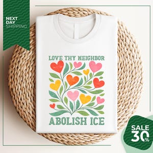 A white t-shirt with a colorful graphic design featuring a tree with heart-shaped leaves and flowers, along with the text "LOVE thy neighbor ABOLISH ICE". The t-shirt is displayed on a woven basket, and there is a green banner with text that reads "NEXT DAY SHIPPING" and "SALE 30%".