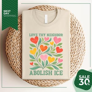 a beige t-shirt with a colorful graphic design of hearts and leaves, along with the text "Love thy neighbor" and "Abolish ice" printed on it. The t-shirt is displayed on a woven basket, and there is a green banner with white text that reads "Next day shipping" and "SALE 30%".