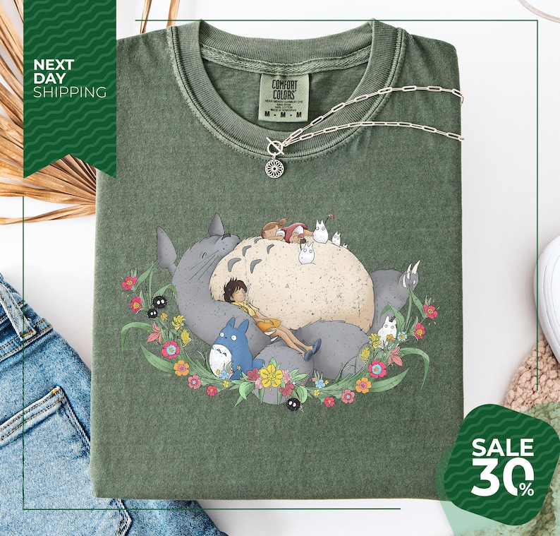 a green t-shirt with a colorful illustration of a cat surrounded by various animals and flowers. The t-shirt is displayed on a white background, and there is a sale sign indicating a 30% discount.
