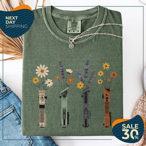 a green t-shirt with a graphic design of four different colored vases holding flowers, arranged in a row. The vases are depicted in various colors, including green, orange, and purple. The t-shirt is displayed on a white background, and there are some jeans and a necklace visible in the background.