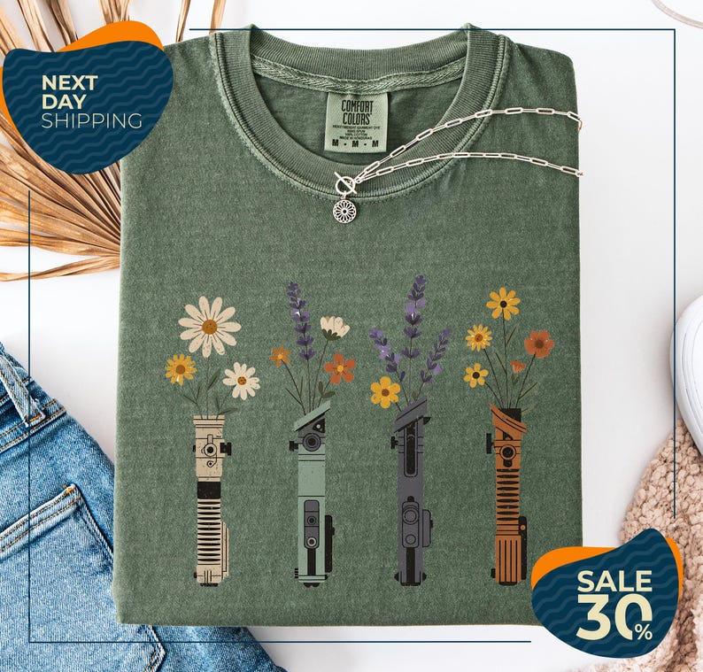 a green t-shirt with a graphic design of four different colored vases holding flowers, arranged in a row. The vases are depicted in various colors, including green, orange, and purple. The t-shirt is displayed on a white background, and there are some jeans and a necklace visible in the background.