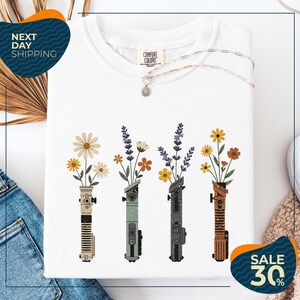 a white t-shirt with a graphic design of four colorful vases holding flowers, arranged in a row. The vases are of different colors and shapes, creating a visually appealing pattern. The t-shirt is displayed on a white background, and there are some jeans and a sweater visible in the background. The image also includes text that reads "Next Day Shipping" and "SALE 30%", indicating that the t-shirt is available for immediate shipping and is on sale at a discounted price.