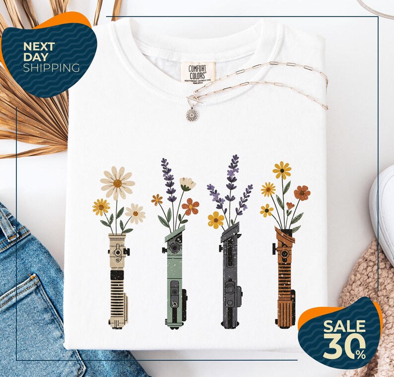 a white t-shirt with a graphic design of four colorful vases holding flowers, arranged in a row. The vases are of different colors and shapes, creating a visually appealing pattern. The t-shirt is displayed on a white background, and there are some jeans and a sweater visible in the background. The image also includes text that reads "Next Day Shipping" and "SALE 30%", indicating that the t-shirt is available for immediate shipping and is on sale at a discounted price.