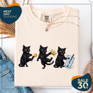 a white t-shirt with a graphic design of three black cats, each holding a different object: a ball, a crown, and a glass of water. The t-shirt is displayed on a white background, and there are some jeans and a sweater visible in the background. The image also includes text that reads "Next Day Shipping" and "SALE 30%".