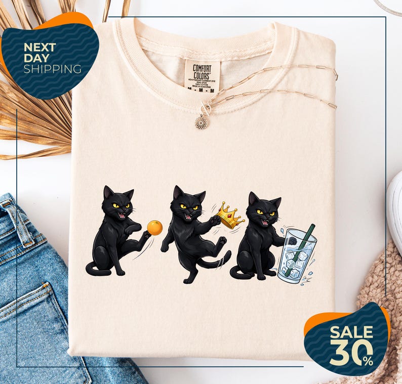 a white t-shirt with a graphic design of three black cats, each holding a different object: a ball, a crown, and a glass of water. The t-shirt is displayed on a white background, and there are some jeans and a sweater visible in the background. The image also includes text that reads "Next Day Shipping" and "SALE 30%".