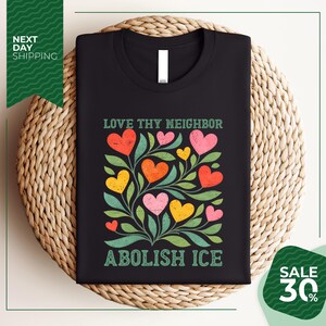 a black t-shirt with a colorful graphic design of hearts and leaves, along with the text "Love thy neighbor" and "Abolish ice" printed on it. The t-shirt is displayed on a woven basket, and there is a sale offer for 30% off.