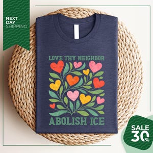 A navy blue t-shirt with a colorful graphic design of hearts and leaves, along with the text "Love thy neighbor, Abolish ice" printed on it. The t-shirt is displayed on a woven placemat, and there is a green banner with text that reads "Next day shipping" and "SALE 30%".