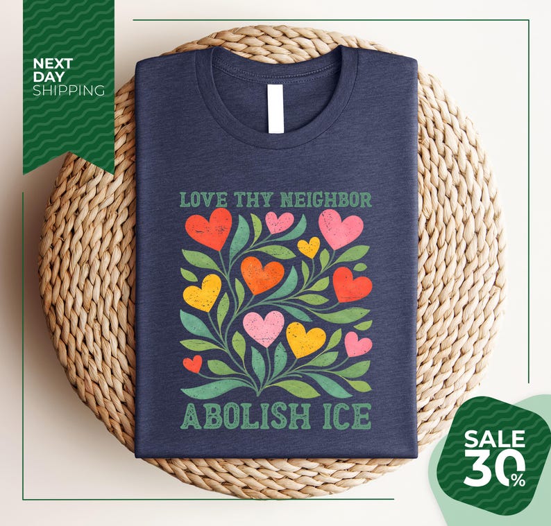 A navy blue t-shirt with a colorful graphic design of hearts and leaves, along with the text "Love thy neighbor, Abolish ice" printed on it. The t-shirt is displayed on a woven placemat, and there is a green banner with text that reads "Next day shipping" and "SALE 30%".